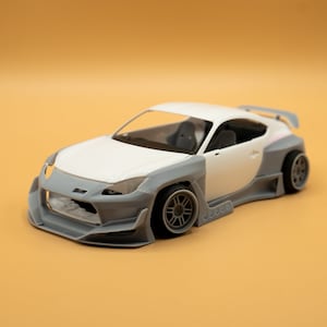 以下が含まれることがあります： 白いとグレーのモデルカー。スポイラーとカスタムボディキットが付いています。車は黒いホイールで、明るいオレンジ色の背景に配置されています。このモデルはスポーツカーの詳細なレプリカです。