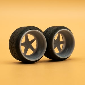 May include: Two black and gray model car wheels with a five-spoke design. The wheels are mounted on a light orange surface.