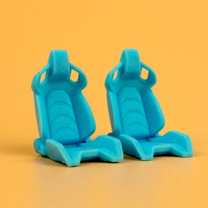May include: Two bright blue racing car seats, made of plastic, are shown against a yellow background. The seats have a sporty design with a high back and side bolsters.