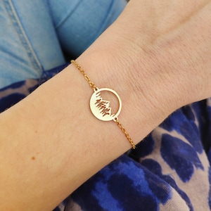 May include: A gold chain bracelet with a circular pendant featuring a cut-out design of a mountain range.