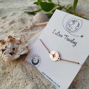 May include: Rose gold compass bracelet with a star in the center and the letters N, S, E, and W around the edge. The bracelet is on a white card with the text "I Love Traveling" and a globe with a plane flying over it. The card also has the website "www.vagabond-life.com" printed on it.
