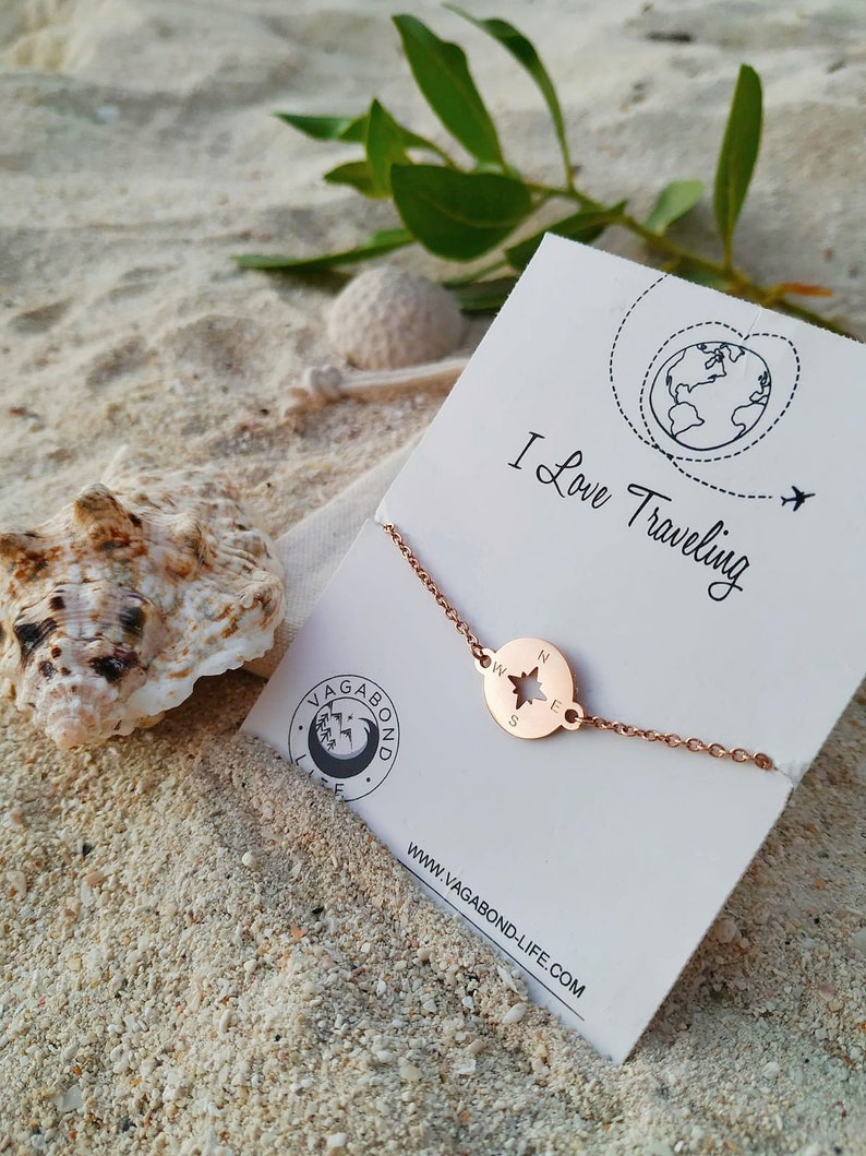 May include: Rose gold compass bracelet with a star in the center and the letters N, S, E, and W around the edge. The bracelet is on a white card with the text "I Love Traveling" and a globe with a plane flying over it. The card also has the website "www.vagabond-life.com" printed on it.