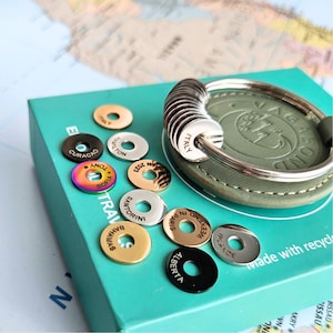 Può includere: Un set di portachiavi rotondi in metallo con nomi di città incisi, tra cui "Curaçao", "Sofia", "Italia", "Santorini", "Amas", "Alberta", "Weekend a Parigi", "Ipiales" e "Weekend a Oslo". I portachiavi sono disposti su una superficie verde con il testo "Realizzato con materiale riciclato" visibile.