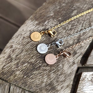 May include: Three gold, silver, and rose gold chains with a circular pendant that reads "Vagabond Life" on each. The chains are on a wooden surface.
