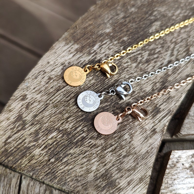 May include: Three gold, silver, and rose gold chains with a circular pendant that reads "Vagabond Life" on each. The chains are on a wooden surface.
