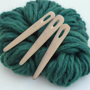 Op de afbeelding: Drie houten garennaalden liggen op een bol donkergroen garen. De naalden zijn lichtbeige met grote oogjes. Het garen is dik en getextureerd, wat een zachte uitstraling geeft. Ideaal voor knutsel- en breiprojecten.