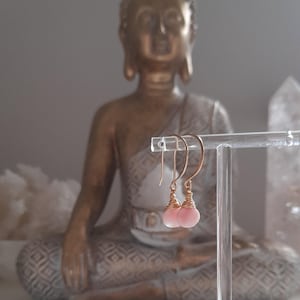 May include: Pink opal earrings with gold-coloured wire wrapping, displayed on a clear stand. The earrings are teardrop-shaped. A gold-coloured Buddha statue is in the background.