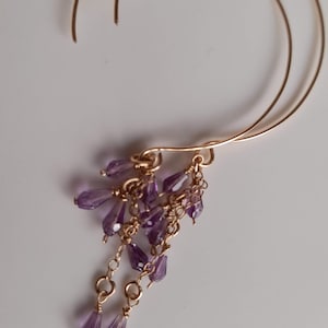 May include: A pair of gold-toned hoop earrings featuring dangling clusters of small, faceted purple teardrop-shaped beads. The earrings are constructed with thin, curved gold-toned wire hoops that suspend multiple delicate chains adorned with purple crystals.