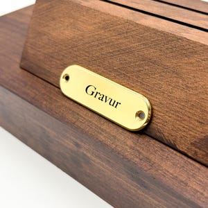 Può includere: Una scatola di legno con una placca di metallo dorato che dice "Gravur".