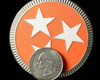 Può includere: Una moneta d'argento con un designo rosso e bianco con tre stelle. La moneta è su una superficie nera e una moneta da un quarto di dollaro d'argento è davanti ad essa.
