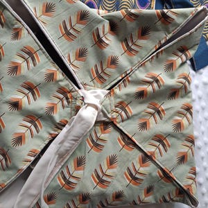May include: A square, quilted fabric item with a feather pattern in shades of brown, orange, and white on a sage green background. The item is tied with a white ribbon. The item is likely a baby blanket or similar textile.