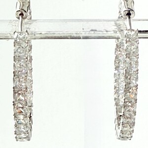 May include: A pair of silver hoop earrings encrusted with clear, sparkling gemstones. The earrings are rectangular in shape and are set in a silver metal. The gemstones are closely set together, creating a continuous line of sparkle.