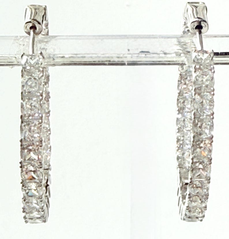 May include: A pair of silver hoop earrings encrusted with clear, sparkling gemstones. The earrings are rectangular in shape and are set in a silver metal. The gemstones are closely set together, creating a continuous line of sparkle.