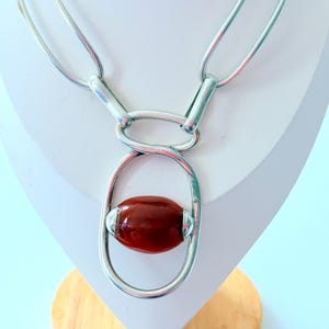 Op de afbeelding: Een zilverkleurige ketting met een grote, ovale, roodbruine stenen hanger. De ketting heeft een schakelkettingontwerp met een langwerpige ovale vorm die de steen vasthoudt. De ketting wordt gepresenteerd op een witte mannequin.