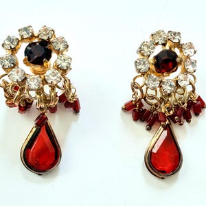 Peut inclure: Une paire de boucles d'oreilles dorées composées d'un cercle de strass transparents entourant une pierre rouge foncé. Sous le cercle, des perles rouges pendent à des maillons dorés, terminées par une grande pierre rouge en forme de goutte.
