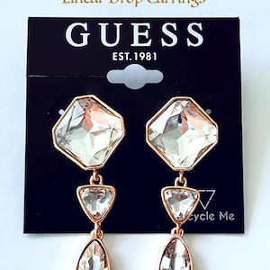 Puede incluir: Pendientes colgantes lineales de cristal Guess de estilo vintage. Los pendientes presentan un cristal grande, transparente y facetado de forma cuadrada en la parte superior, con un triángulo más pequeño y un cristal de forma ovalada debajo. El metal es de color oro rosa. El logotipo de Guess es visible.
