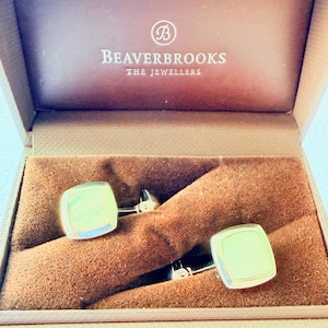 May include: A pair of silver-toned square cufflinks with a smooth, reflective surface, presented inside a brown velvet-lined box. The interior lid of the box features the white printed text "B BEAVERBROOKS THE JEWELLERS" centered at the top.