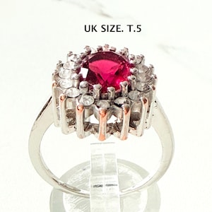 Op de afbeelding: Zilverkleurige ring met een grote, ronde, robijnrode edelsteen in het midden, omringd door kleinere, heldere stenen. De ring heeft een halo-ontwerp en maat UK T.5.