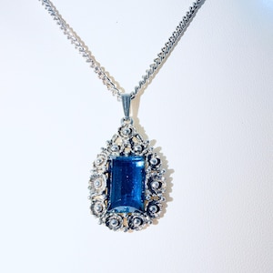 Op de afbeelding: Een zilverkleurige ketting met een rechthoekige, diepblauwe gefacetteerde edelsteen hanger. De hanger is omgeven door een ingewikkeld, sierlijk metalen frame met kleine cirkelvormige details, hangend aan een bijpassende zilverkleurige metalen ketting.