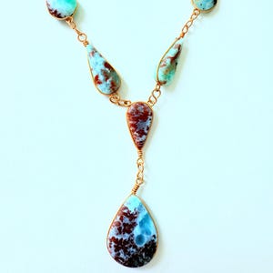 May include: A handcrafted necklace featuring teardrop-shaped gemstones in shades of blue, brown, and white. The stones are connected by delicate copper-colored wire and chain, creating a Y-shaped pendant. The necklace is displayed against a white background.