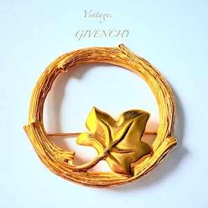 May include: Vintage gold-tone Givenchy brooch. The brooch features a circular design with a textured, branch-like frame and a gold leaf accent. The word "Vintage" and the brand name "GIVENCHY" are printed above the brooch.