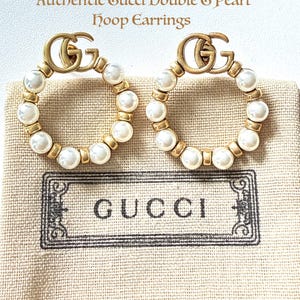 Puede incluir: Pendientes de aro Gucci Double G dorados con perlas de imitación. Los pendientes presentan el logotipo de Gucci en la parte superior y se muestran sobre una tela beige con el logotipo de Gucci impreso. El texto "Authentic Gucci Double G Pearl Hoop Earrings" está en la parte superior.