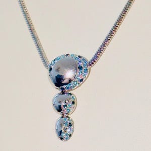 Pode incluir: Colar prateado com um pendente composto por três elementos ovais de tamanho decrescente. O elemento superior é um círculo maior, todos adornados com pequenas pedras preciosas coloridas. A corrente tem um design fino e serpentino.