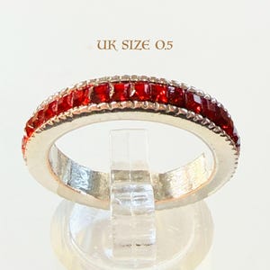 May include: A silver ring with a band of square-cut, vibrant red gemstones. The ring is set against a white background and the text "UK SIZE 0.5" is visible above the ring.