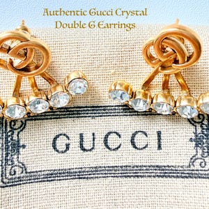 Puede incluir: Un par de pendientes Gucci Double G dorados con detalles de cristal. Cada pendiente presenta el icónico logotipo G entrelazado y cinco cristales transparentes. Los pendientes se muestran sobre una tela beige con el logotipo de Gucci.
