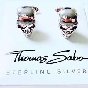 May include: A pair of sterling silver skull cufflinks. The cufflinks have a polished finish and are presented on a white card with the brand name "Thomas Sabo" and the words "STERLING SILVER".