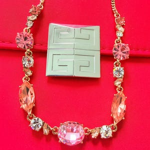 May include: A gold-tone chain necklace featuring a series of pink and clear faceted crystals in round and marquise shapes. Positioned above the necklace is a square silver-tone metal plaque engraved with the Givenchy logo in a four-part design.