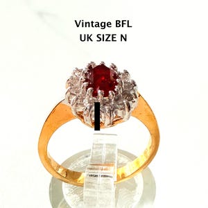 以下が含まれることがあります： 大きな楕円形の濃い赤色の宝石を小さな透明な石で囲んだ、ヴィンテージのゴールドリング。 リングはゴールドバンドで、透明なスタンドに展示されています。 画像には「Vintage BFL UK SIZE N」というテキストが含まれています。