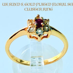 May include: Gold-plated floral cluster ring featuring a central ruby and surrounding clear and teal-colored stones. The ring has a delicate band and a floral design. The image text reads "UK SIZED S. GOLD PLATED FLORAL SET CLUSTER RING."