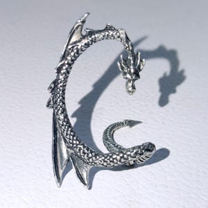 May include: A silver-toned ear cuff shaped like a dragon with a textured, scale-like body that curves along the ear. The dragon head features detailed facial features, while the tail curls inward at the bottom, and fin-like structures extend from the back.