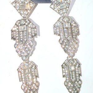 May include: A pair of long, silver-toned drop earrings featuring a geometric, multi-tiered design. Each earring is encrusted with numerous clear, sparkling rhinestones and baguette-cut crystals arranged in a symmetrical, intricate pattern.
