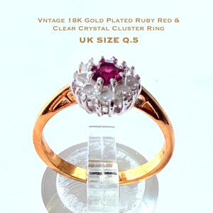 以下が含まれることがあります： ヴィンテージ18K金メッキリング。クリアクリスタルのクラスターと中央にルビーレッドの石が特徴です。リングはゴールドバンドで、UKサイズQ.5です。リングは透明なスタンドに展示されています。
