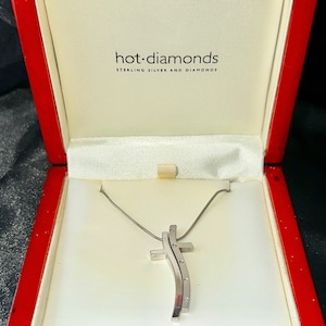 Puede incluir: Un collar con colgante de cruz plateada expuesto en una caja roja. La cruz tiene un diseño curvo con pequeños detalles brillantes. La caja está forrada con tela blanca y tiene el texto "hot diamonds STERLING SILVER AND DIAMONDS" impreso en el interior.