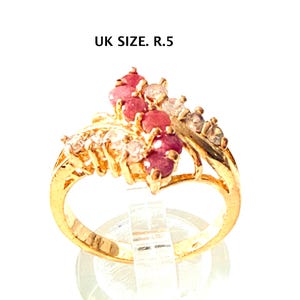 May include: A gold ring set with red and clear gemstones. The ring has a cluster design and a split shank. The text "UK SIZE. R.5" is visible.