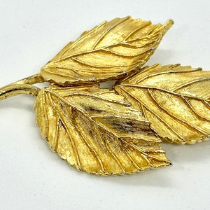 May include: A gold-toned leaf brooch with three leaves, each with a detailed vein pattern. The leaves are arranged in a cluster, with the stem extending to the side.