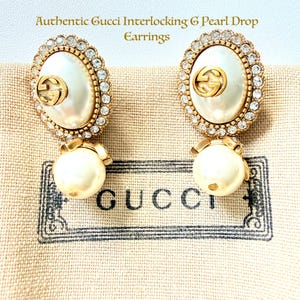 May include: Pair of gold-toned Gucci earrings with an oval-shaped pearl center, surrounded by clear crystals. Each earring features a gold Gucci logo and a dangling pearl. The text at the top reads "Authentic Gucci Interlocking G Pearl Drop Earrings."