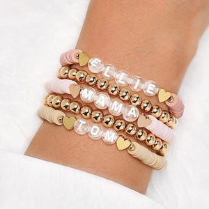 May include: Stack of beaded bracelets in shades of pink and beige, with gold accents. Each bracelet features a different name spelled out in white letters. Gold heart charms and gold beads add detail to the bracelets.