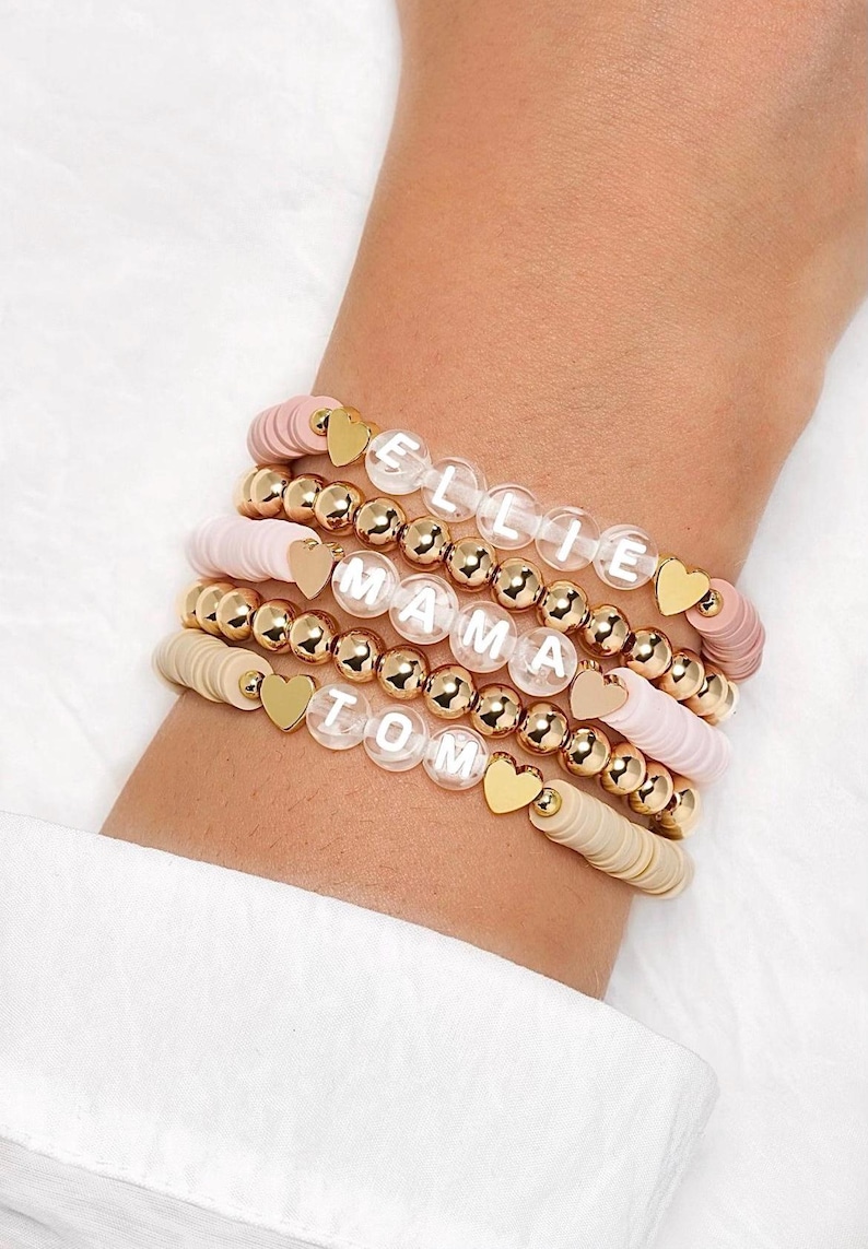 May include: Stack of beaded bracelets in shades of pink and beige, with gold accents. Each bracelet features a different name spelled out in white letters. Gold heart charms and gold beads add detail to the bracelets.