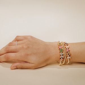 May include: A hand wearing a silver ring and three gold bracelets. The bracelets spell out "DANCE" and "GRACE" with colourful letter beads. The bracelets are on a light beige background.