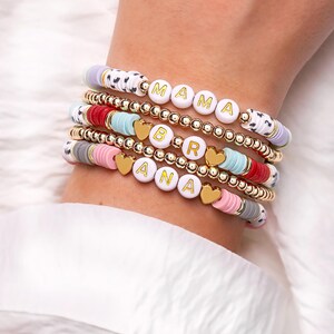 May include: Stack of beaded bracelets with white letter beads spelling "MAMA" and "BRANA". The bracelets feature gold beads, heart charms, and colorful disc beads in pink, red, blue, and gray. The bracelets are on a wrist.