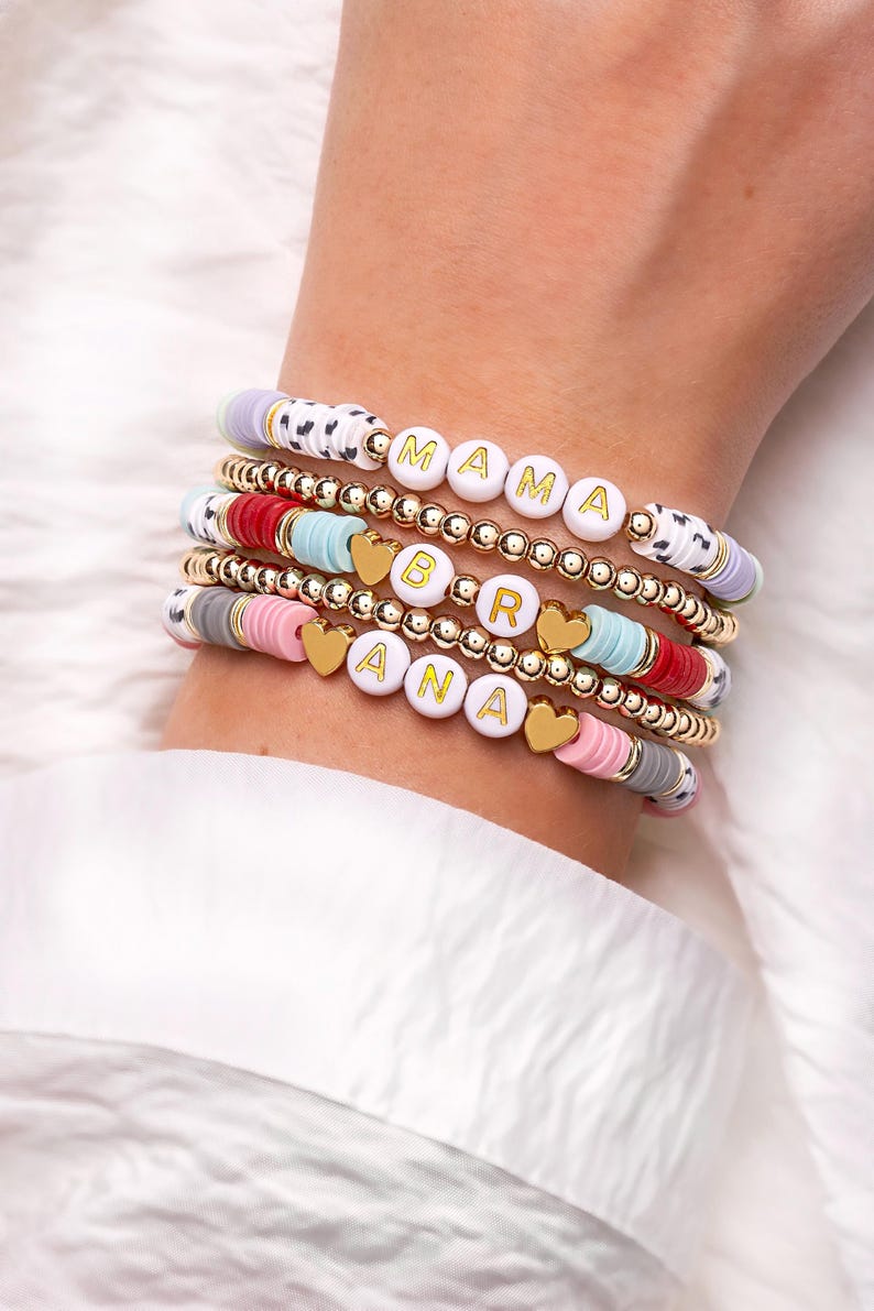 May include: Stack of beaded bracelets with white letter beads spelling "MAMA" and "BRANA". The bracelets feature gold beads, heart charms, and colorful disc beads in pink, red, blue, and gray. The bracelets are on a wrist.