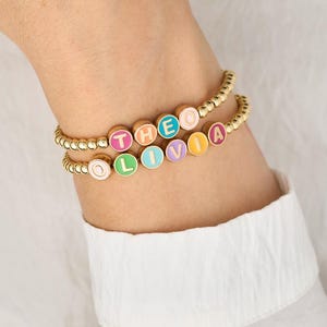 May include: Gold beaded bracelets with colourful enamel letter beads spelling "THEO" and "OLIVIA". The bracelets are worn on a wrist, with a white shirt in the background. The letters are in various colours.