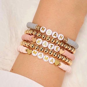 May include: A collection of personalised bracelets. The bracelets feature white letter beads, gold beads, and colourful flat beads. The bracelets spell out "NOAH", "KATE", and "MAMA". Gold heart charms are also included.
