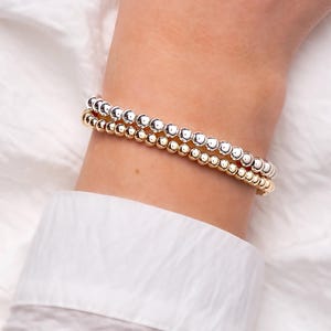 May include: Two beaded bracelets, one gold and one silver, are stacked on a wrist. The bracelets are made of small, round beads. The wrist is against a white background, and the person is wearing a white shirt.