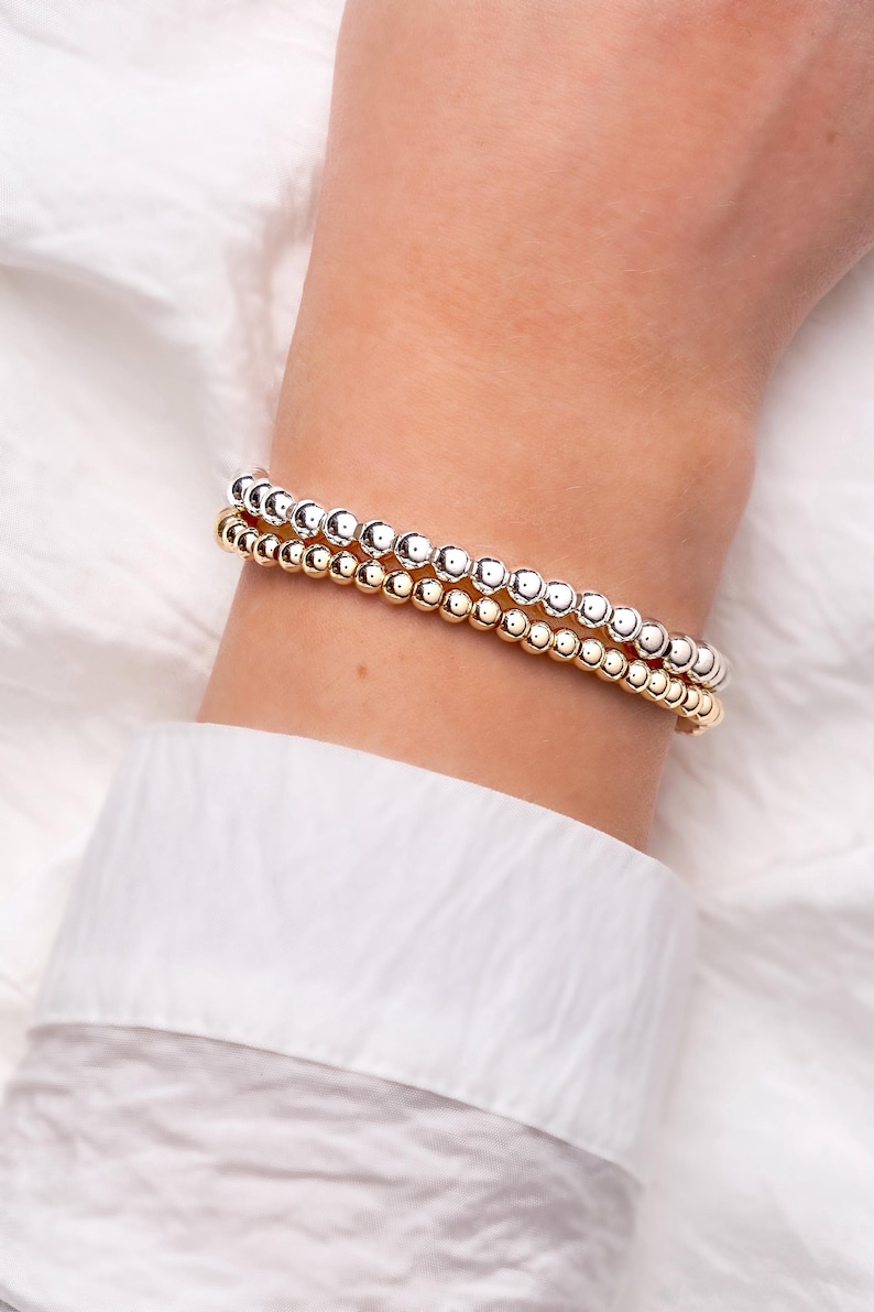 May include: Two beaded bracelets, one gold and one silver, are stacked on a wrist. The bracelets are made of small, round beads. The wrist is against a white background, and the person is wearing a white shirt.