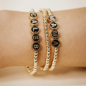 May include: Three gold beaded bracelets. Two bracelets spell out "CALEB" and "MAMA" with black letters on gold circular beads. The third bracelet is made of small gold beads. The bracelets are on a person's wrist.